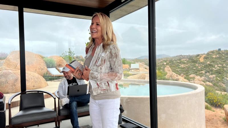 Photo of a happy woman speaking while standing in front of large glass windows with a view to the exterior grounds of a desert resort.