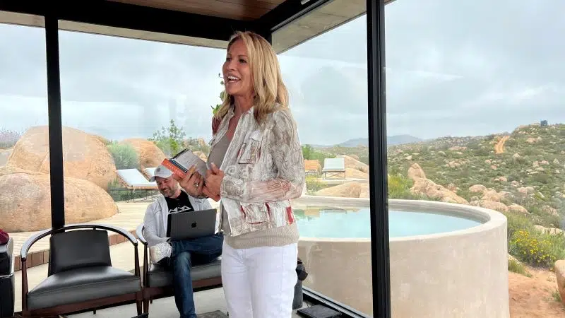 Photo of a happy woman speaking while standing in front of large glass windows with a view to the exterior grounds of a desert resort.